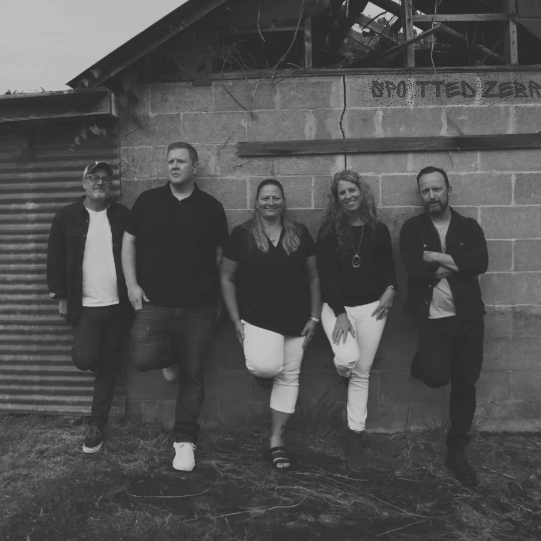 The five members of Spotted Zebra leaning against a weathered cinder-block wall with the band name painted on it